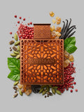 A square fragrance bottle with an ornate geometric cutout design and the brand name RAYHAAN Vantage on the front, styled on a light gray background with botanical and spice props including green leaves, red berries, vanilla pods, and candied fruit.